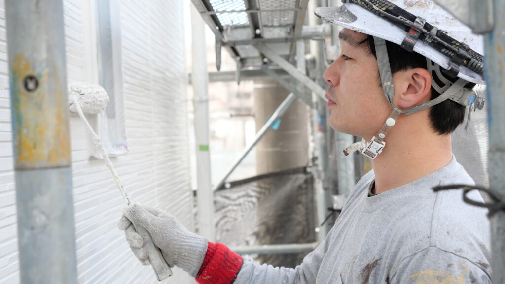 ヘルメットを着用した職人が、足場で白い外壁にローラー塗装をする様子。